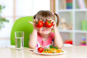 Kleines Mädchen sitzt am Tisch, vor ihr eine Teller mit Essen und hält 2 Tomaten vor die Augen.