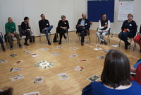 Workshopteilnehmerinnen und -teilnehmer sitzen im Sesselkreis. Am Boden sind in der Mitte des Sesselkreises Fotos aufgelegt, die die Umsetzung der Maßnahme veranschaulichen.