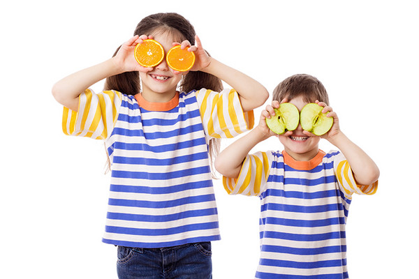 Lächelndes Mädchen hält sich Orangen vor die Augen. Lächelnder Bub hält sich Apfelhälften vor die Augen.
