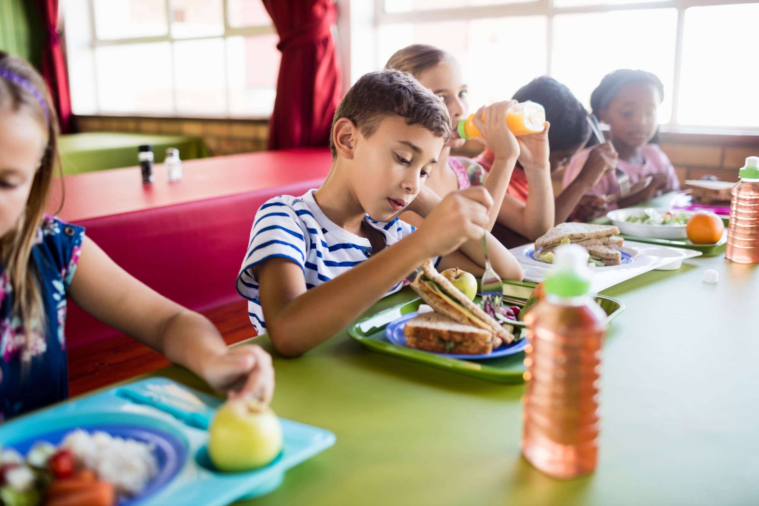 Kinder essen gemeinsam eine Mahlzeit mit Sandwiches, Obst und Getränken in einer hellen Schulkantine.